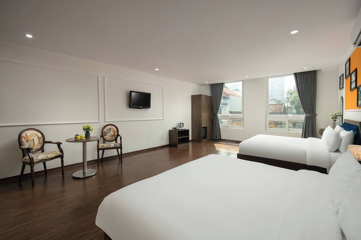A hotel room with a large bed, two armchairs, a small table with a vase, a flat-screen TV mounted on the wall, and a window with curtains.