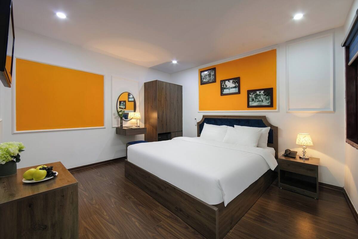 A modern hotel room with a large bed, wooden flooring, a wardrobe, a desk with fruit, and wall-mounted artwork.