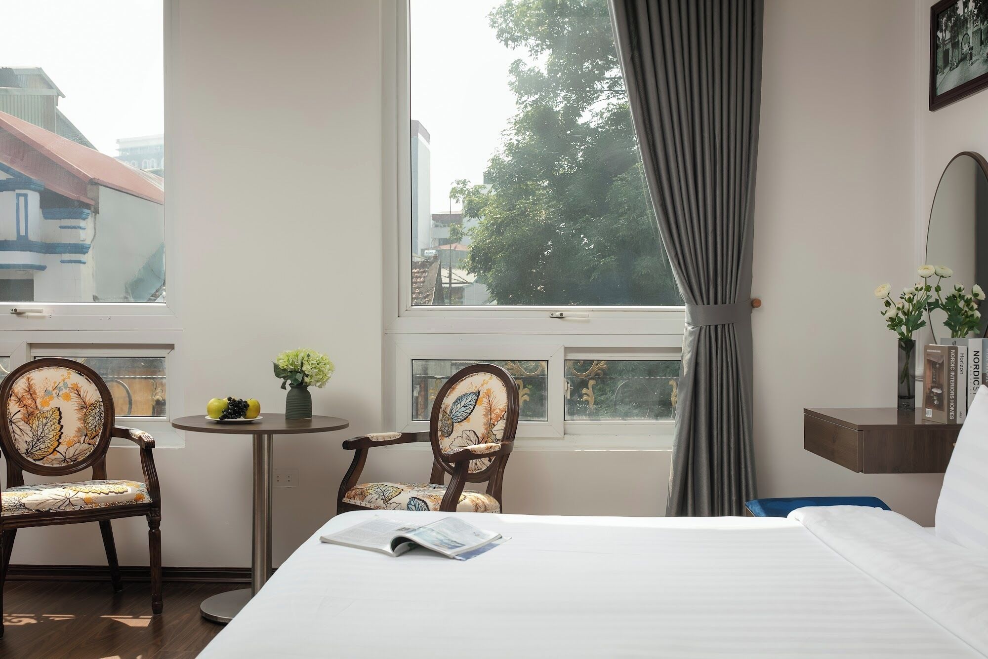 Et hotelværelse med seng, to stole, et lille bord med en vase og frugt, et vindue med gardiner og udsigt til bygninger og træer.