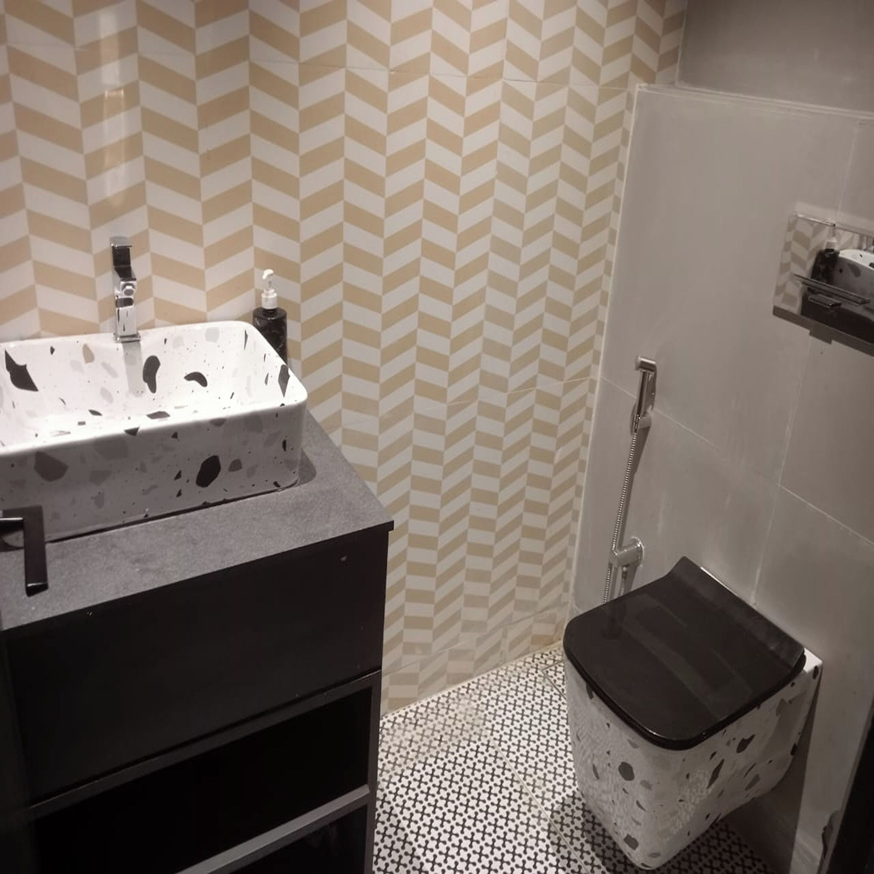 Un baño con un lavamanos blanco, un mueble negro, una ducha con puerta blanca y un suelo estampado.