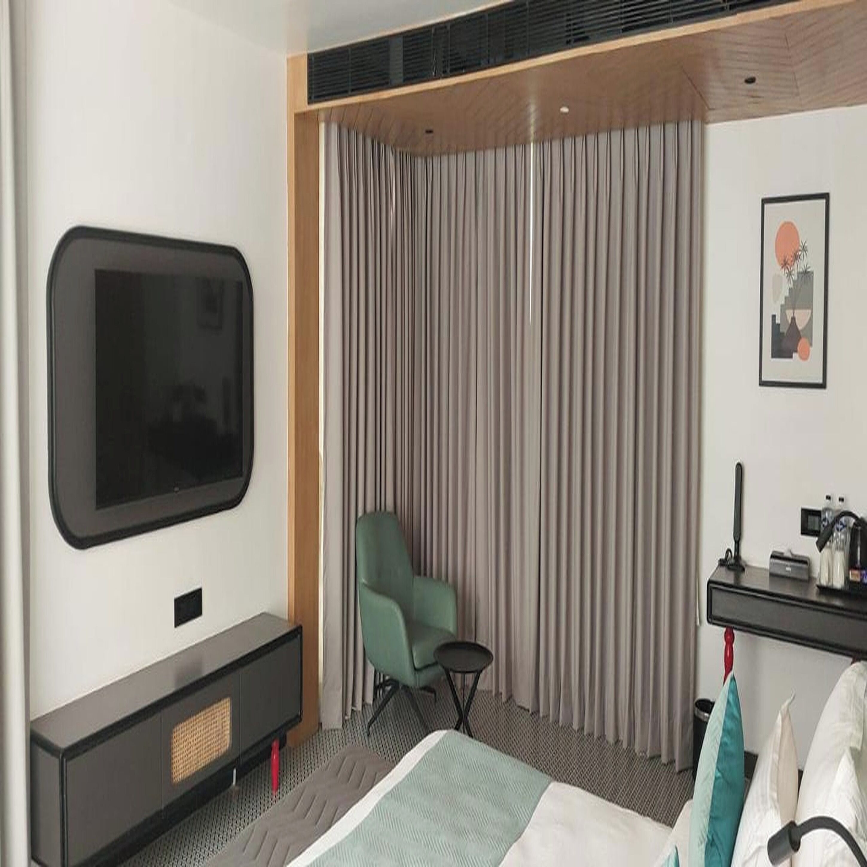 Habitación de hotel con una televisión de pantalla plana montada en la pared, un sillón verde, una cama con colcha blanca y azul turquesa, y una mesita de noche con una cafetera y otros objetos.