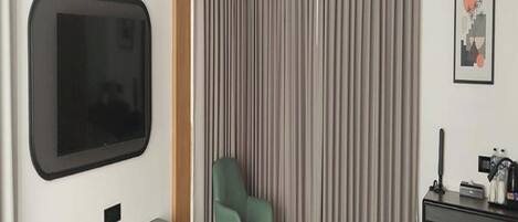 Habitación de hotel con una televisión de pantalla plana montada en la pared, un sillón verde, una cama con colcha blanca y azul turquesa, y una mesita de noche con una cafetera y otros objetos.