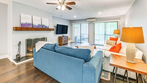 A modern living room with a blue sofa, a wooden side table with a lamp, a ceiling fan, a fireplace, and a large window with blinds. - Sapphire Valley Condo w/ Pool & Lake Beach Access! (Sapphire)