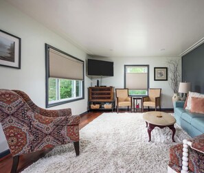 Una sala de estar con un sofá estampado, un sofá azul, una alfombra de pelo blanco, un armario de madera, un televisor y dos sillones.