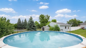 A swimming pool with a wooden deck, surrounded by a fence, trees, and a small shed.