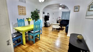 Un área de comedor con piso de madera, una mesa amarilla y sillas azules. Hay un ventilador de techo, un televisor montado en la pared y una puerta con ventana.