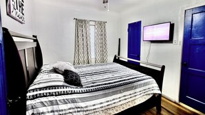 Un dormitorio con cama, ventilador de techo, televisión y ventana con cortinas estampadas.