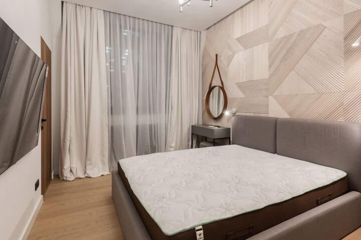 Une chambre à coucher moderne avec un lit, une table de chevet, un miroir suspendu et un mur orné d’un motif géométrique.