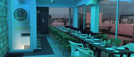 A restaurant with a long dining table, chairs, and a view through large windows.