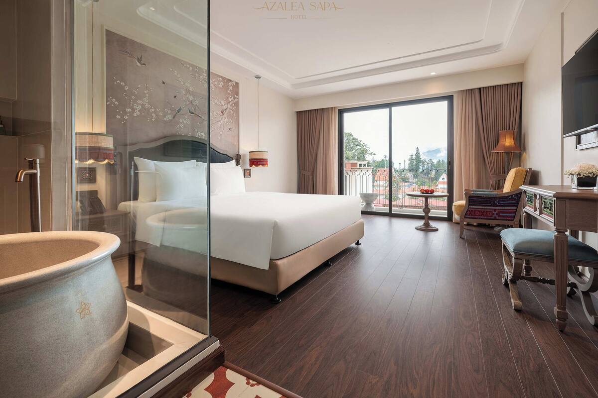 A hotel room with a large bed, a glass partition, a view of the city, and a dining area with a table and chairs.