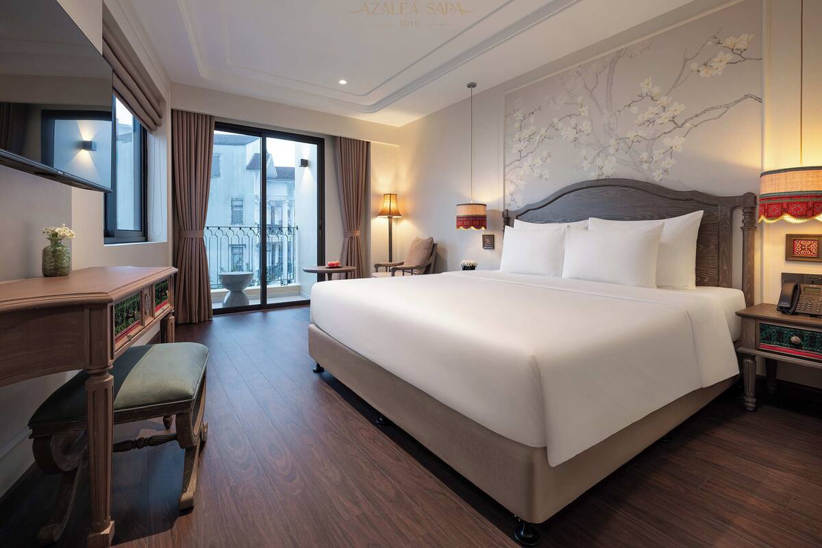 A hotel room with a large bed, a desk, a chair, a small table, and a balcony with curtains.