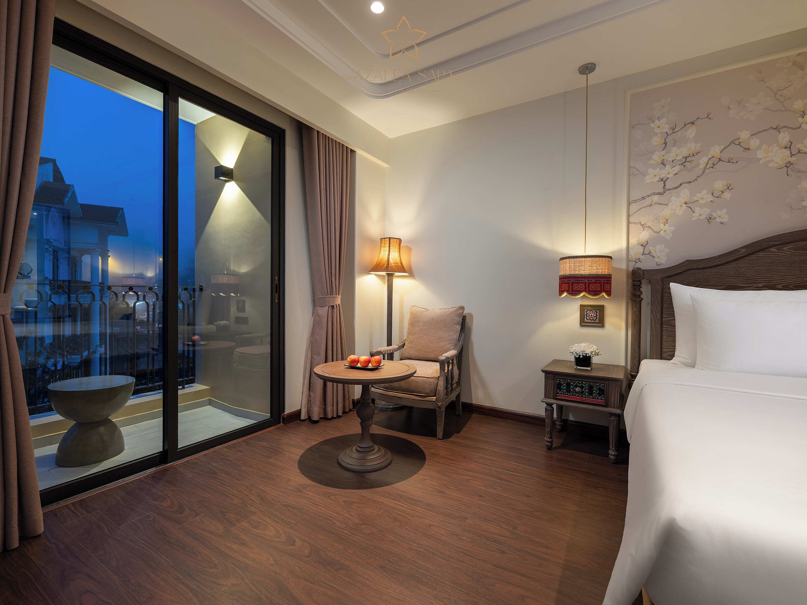 A hotel room with a bed, a chair, a small table with fruit, a lamp, and a view of a building through a glass door.