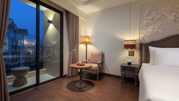 A hotel room with a bed, a chair, a small table with fruit, a lamp, and a view of a building through a glass door.