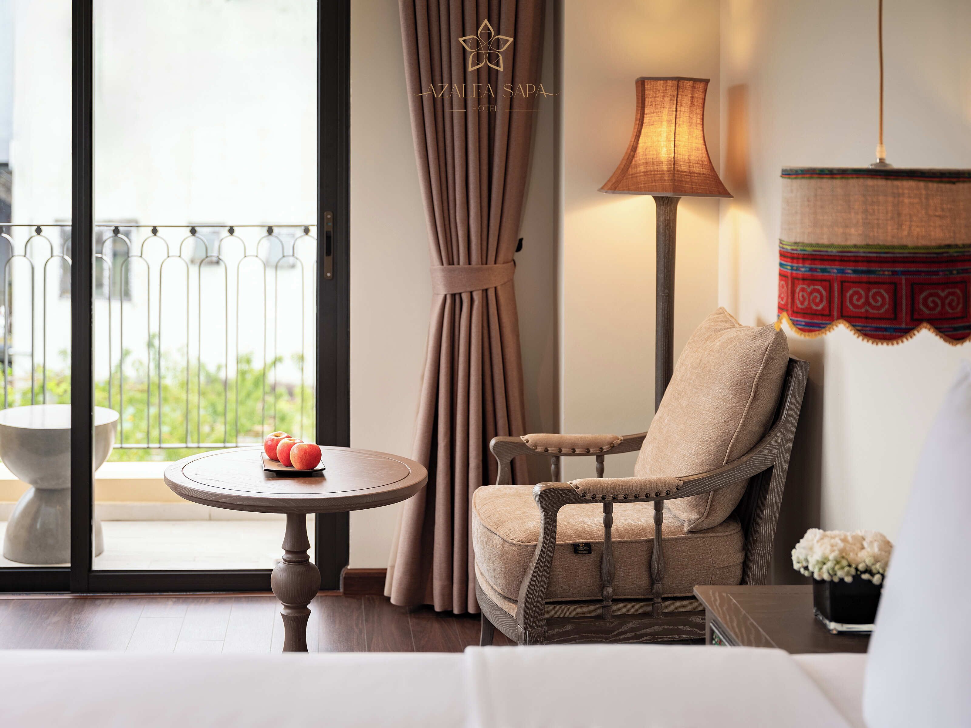 A hotel room with a view of greenery, a round table with apples, a sofa, a lamp, and a decorative hanging.