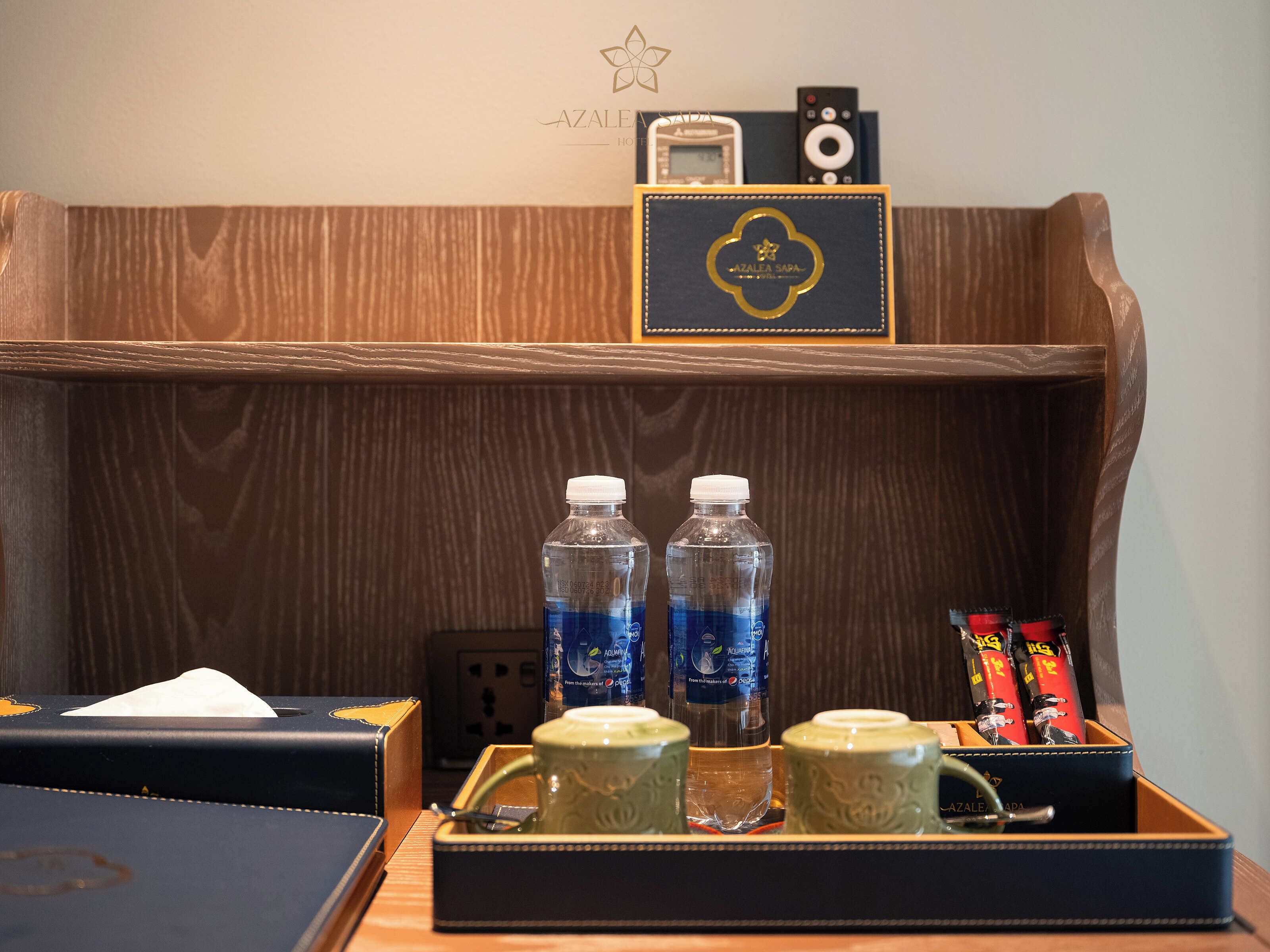 A hotel reception desk with a wooden backdrop, a black box with 'AZALEA SARA' branding, two water bottles, a tray with green mugs, and a remote control.