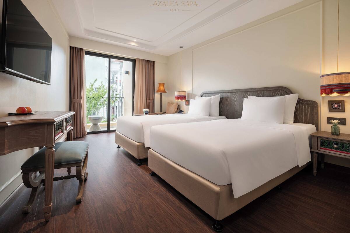 A hotel room with two beds, a flat-screen TV, a desk, and a balcony with curtains.