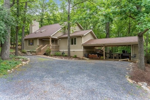 A two-story house with a covered porch, a gravel driveway, and a forested backdrop.