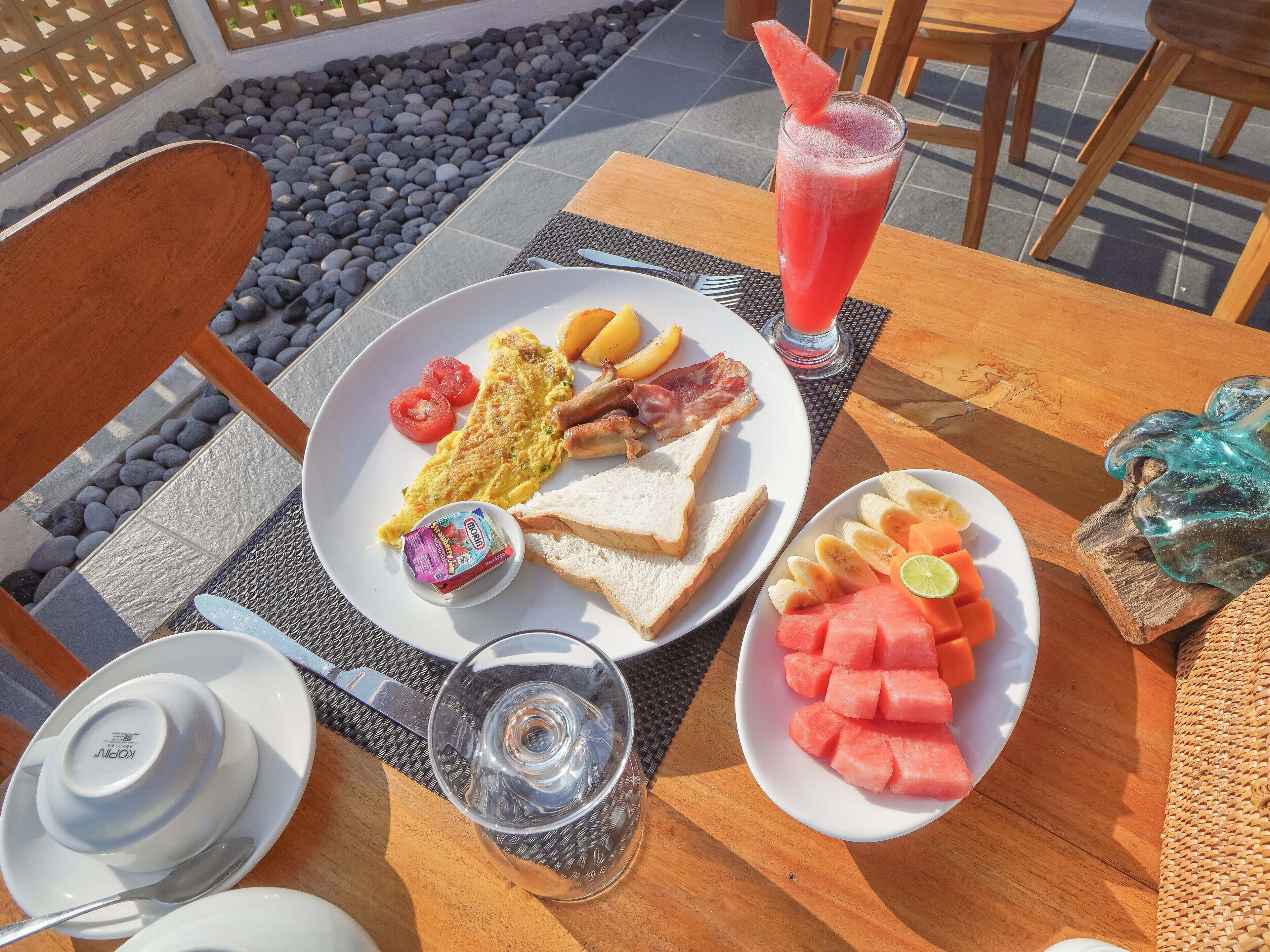 A breakfast setting with a plate of eggs, bacon, toast, tomatoes, and a glass of juice on an outdoor table.