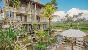 A two-story building with a thatched roof, surrounded by a garden with a small pond, palm trees, and stone pathways. - The Nenggala Suite (Taro)