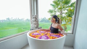 A person sitting in a bathtub filled with colorful flowers, with a view of a green landscape through a window. - The Nenggala Suite (Taro)