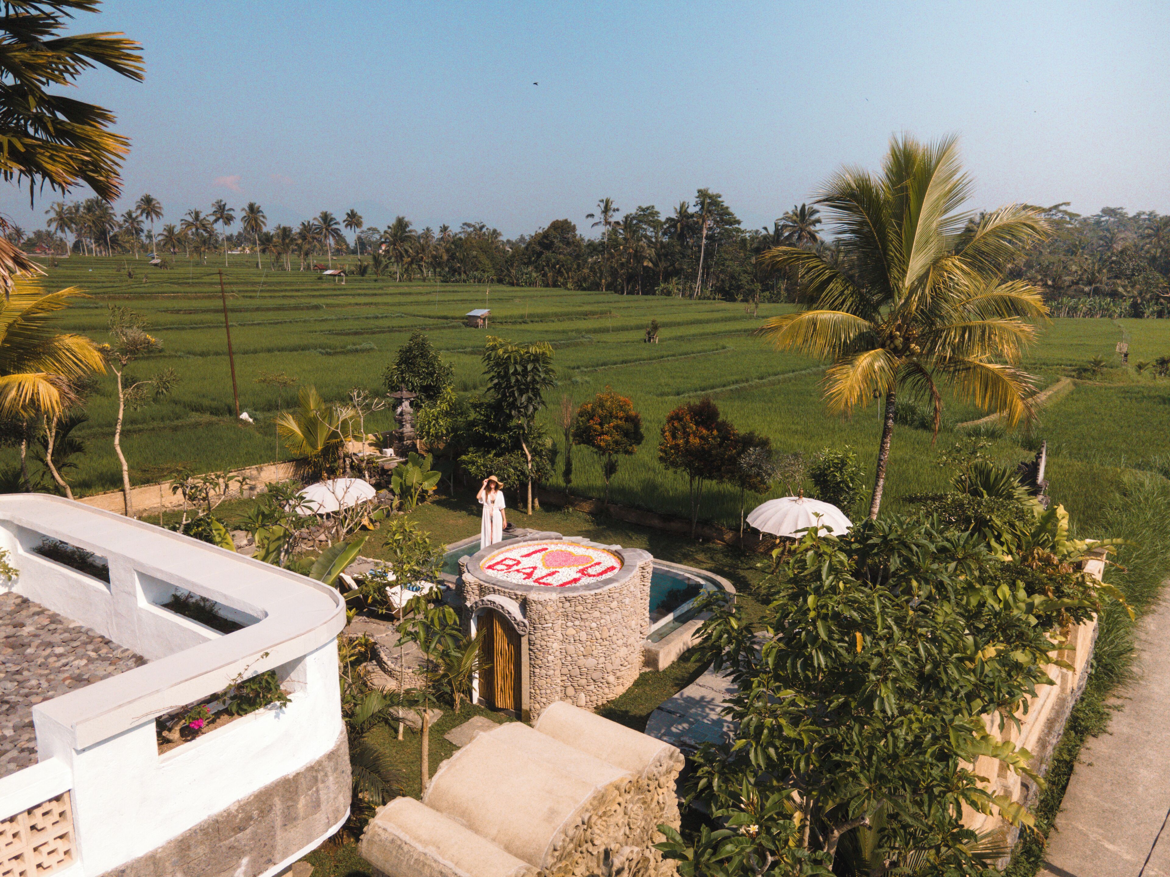 A view of a lush green field with palm trees, a stone structure with a circular design, and a white building with a balcony.