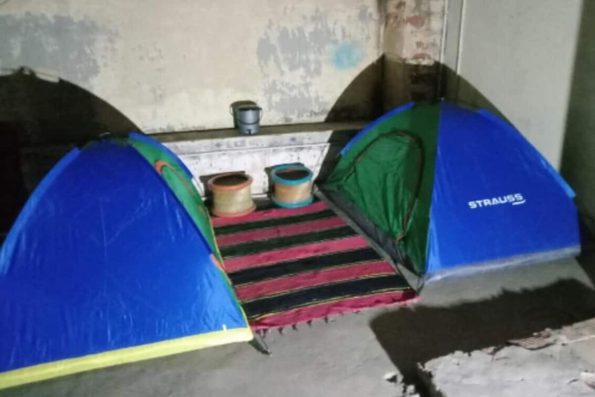 Een kamer met twee blauwe en groene tenten, gelabeld 'STRAUSS', op de vloer, een gestreept kleed en een kleine tafel met een waterkoker en kopjes.