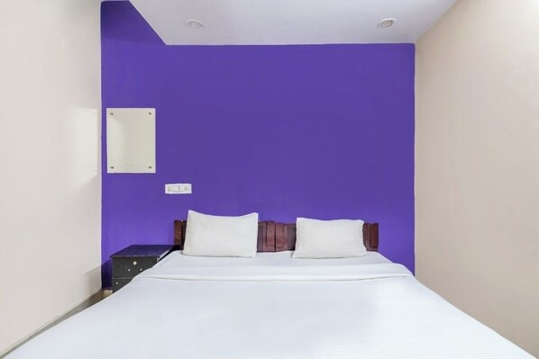 Ein Zimmer mit einem Einzelbett, weißer Bettwäsche, einer violetten Wand und einem kleinen Nachttisch mit einer Lampe.