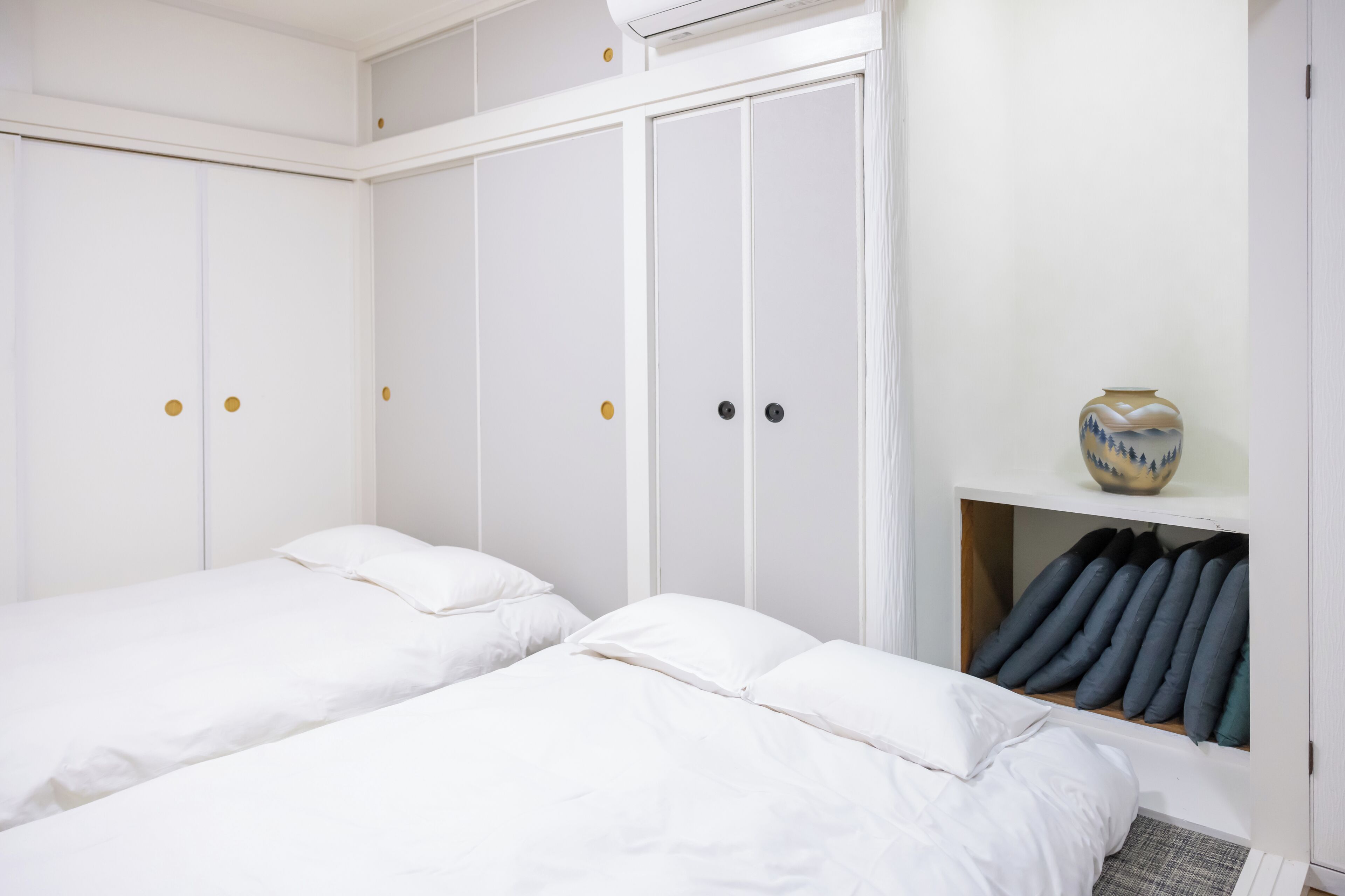 A bedroom with two single beds, white walls, a wardrobe with multiple doors, and a shelf with pillows and a decorative vase.