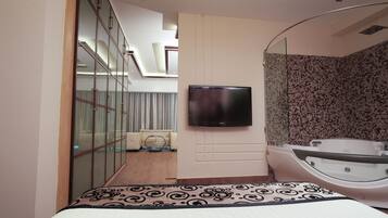 A modern hotel room with a large bed, a TV mounted on the wall, a separate bathroom with a bathtub, and a glass partition separating the room from the hallway.