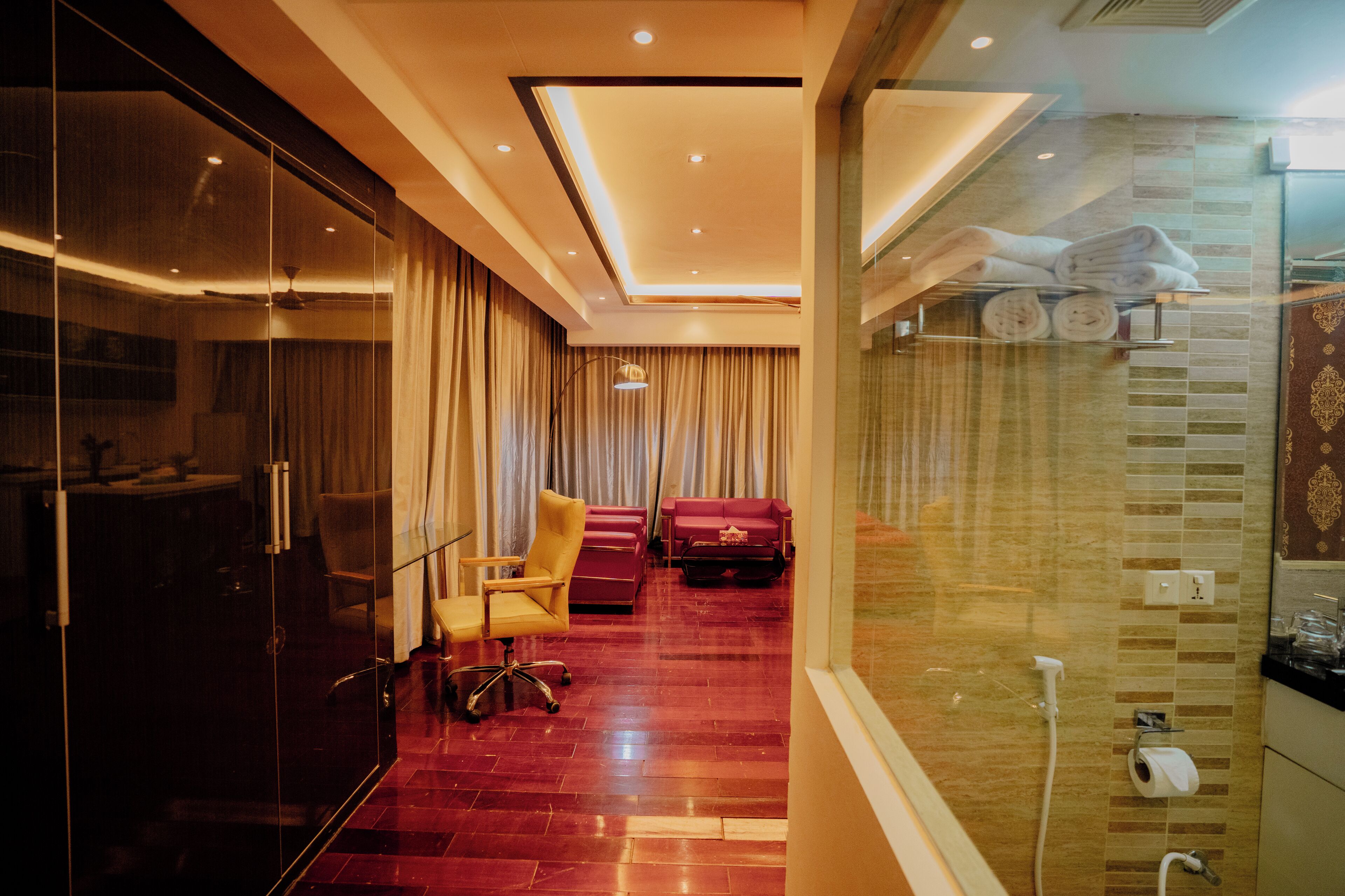 A hotel room with a glass partition, a red carpeted floor, a seating area with a chair and sofa, and a shower area with towels.