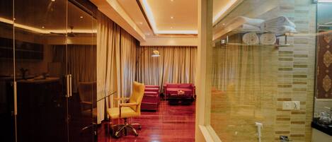 A hotel room with a glass partition, a red carpeted floor, a seating area with a chair and sofa, and a shower area with towels.