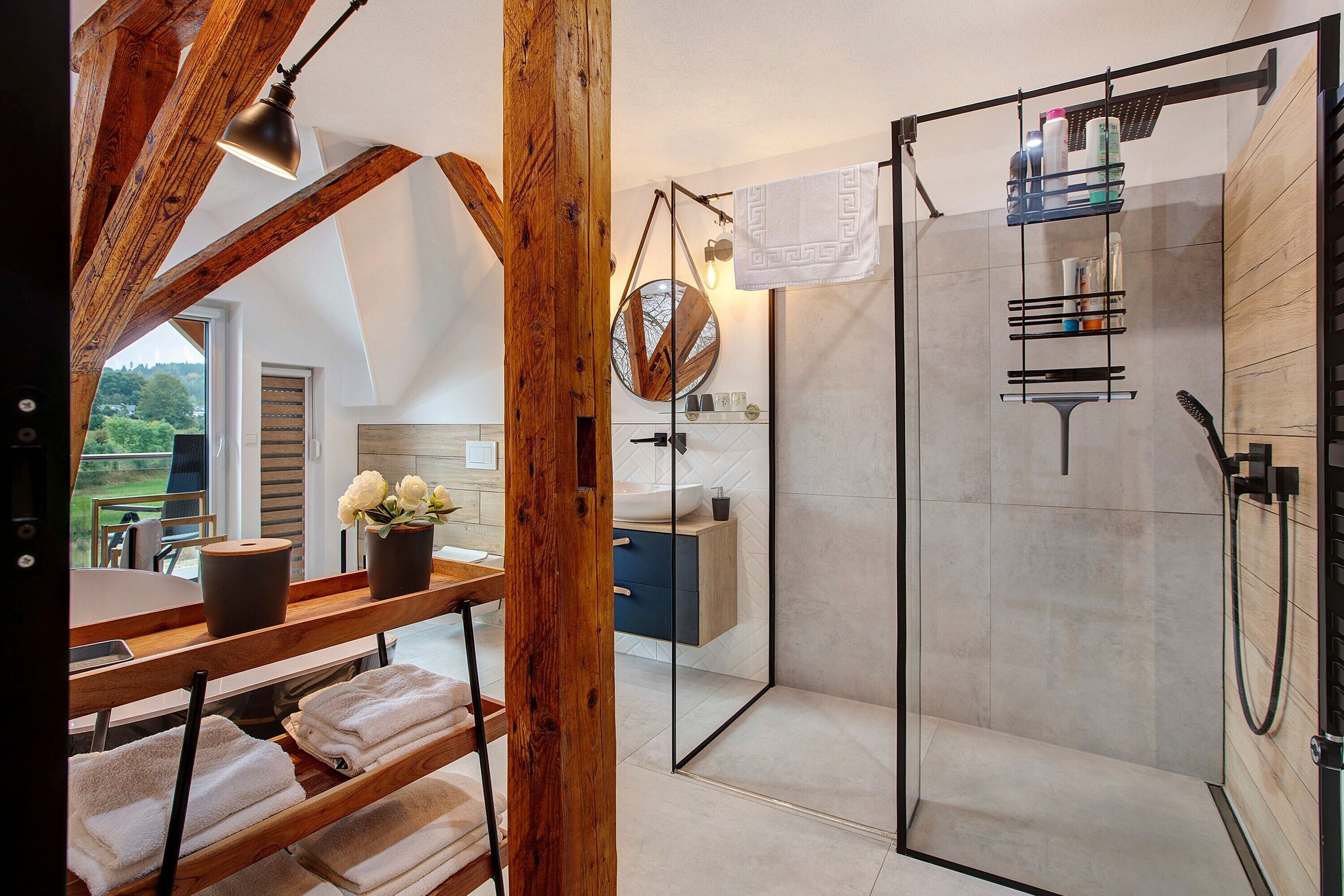 A modern bathroom with a glass shower enclosure, wooden beams, and a view of the outdoors.