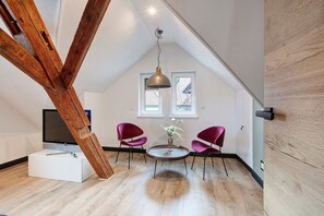 A loft-style room with exposed wooden beams, a round table with chairs, a TV, and a vase with flowers.