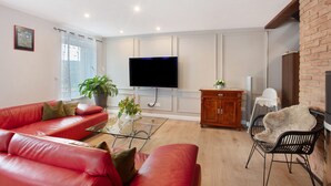 A living room with a red leather sofa, a glass coffee table, a TV, a wooden cabinet, and a brick wall.
