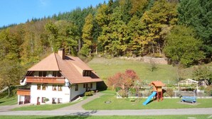 Une maison à deux étages, avec un toit recouvert de tuiles rouges, un balcon et un petit jardin agrémenté d’un toboggan.