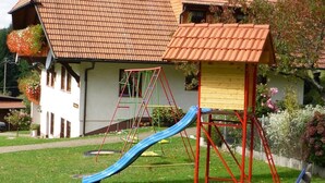 Une maison avec un toit recouvert de tuiles rouges, un toboggan bleu et un banc en bois.