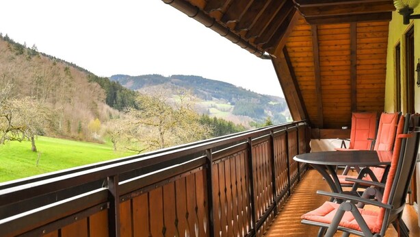 Un balcon en bois, agrémenté d’une table et de chaises, qui offre une vue sur un paysage verdoyant, avec des arbres et des collines.