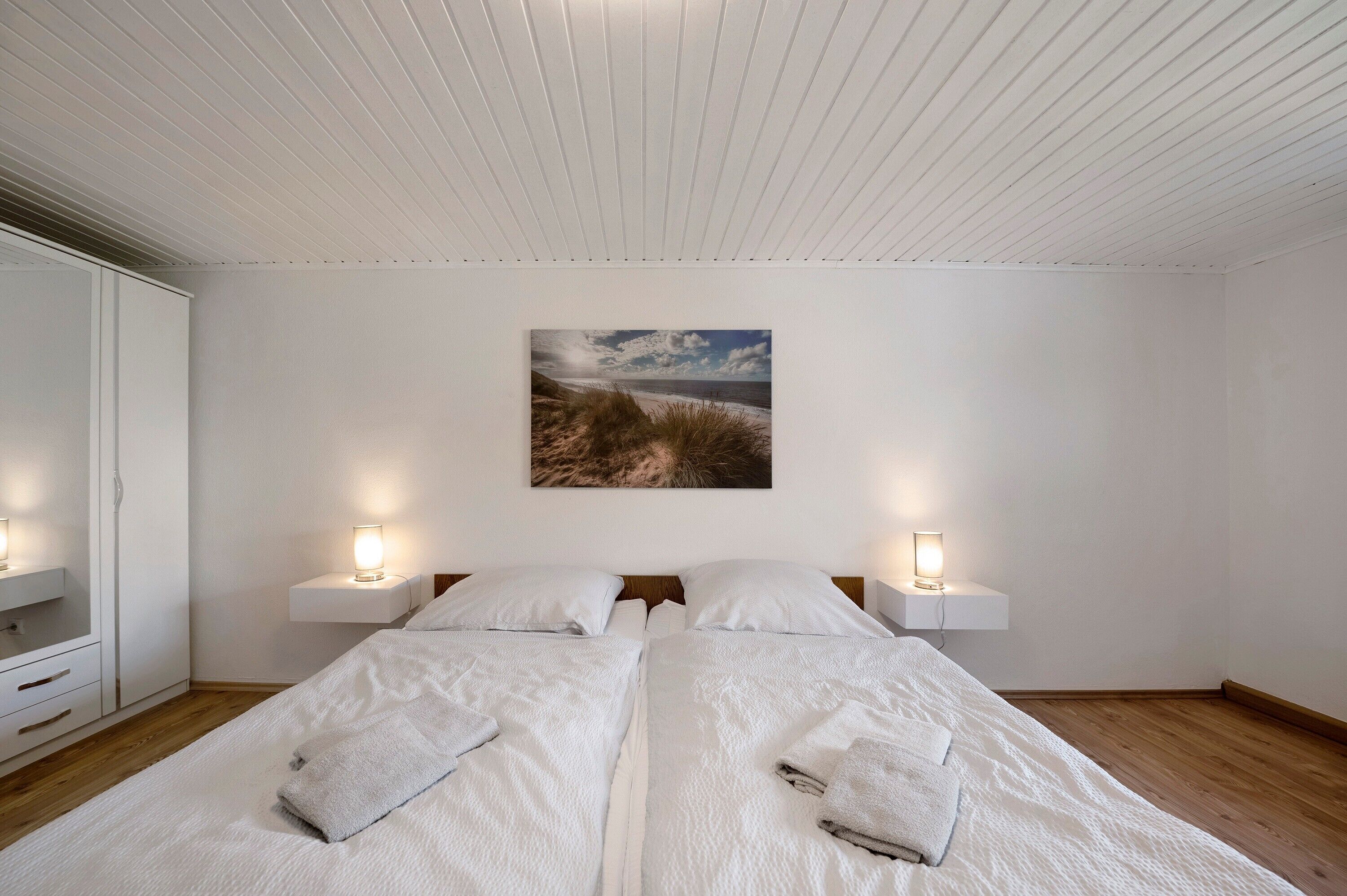 Ein Schlafzimmer mit zwei Betten, einem weißen Kleiderschrank, Nachttischlampen und einem Gemälde einer Strandlandschaft an der Wand.