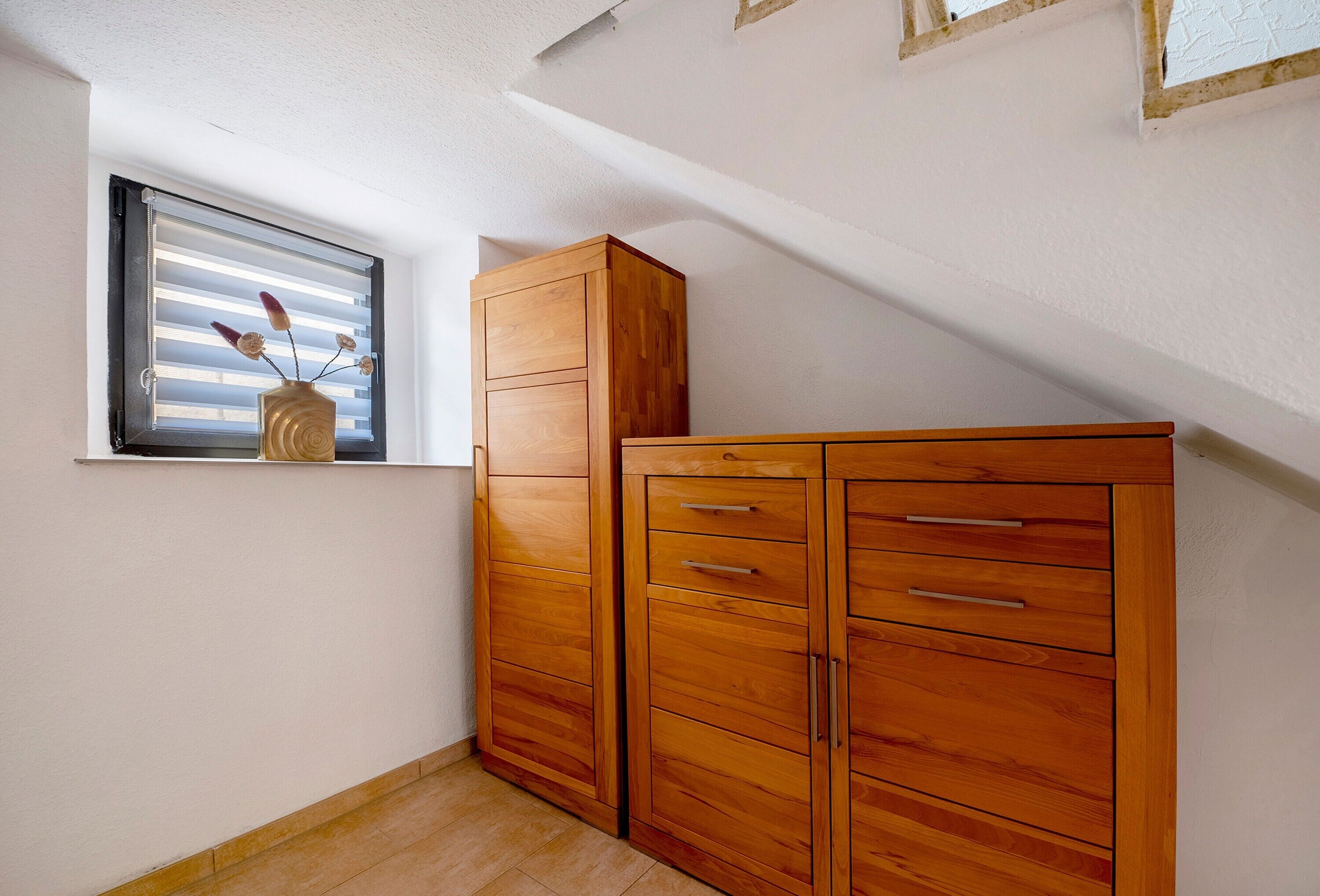A room with a wooden wardrobe, a window with blinds, and a vase with flowers.