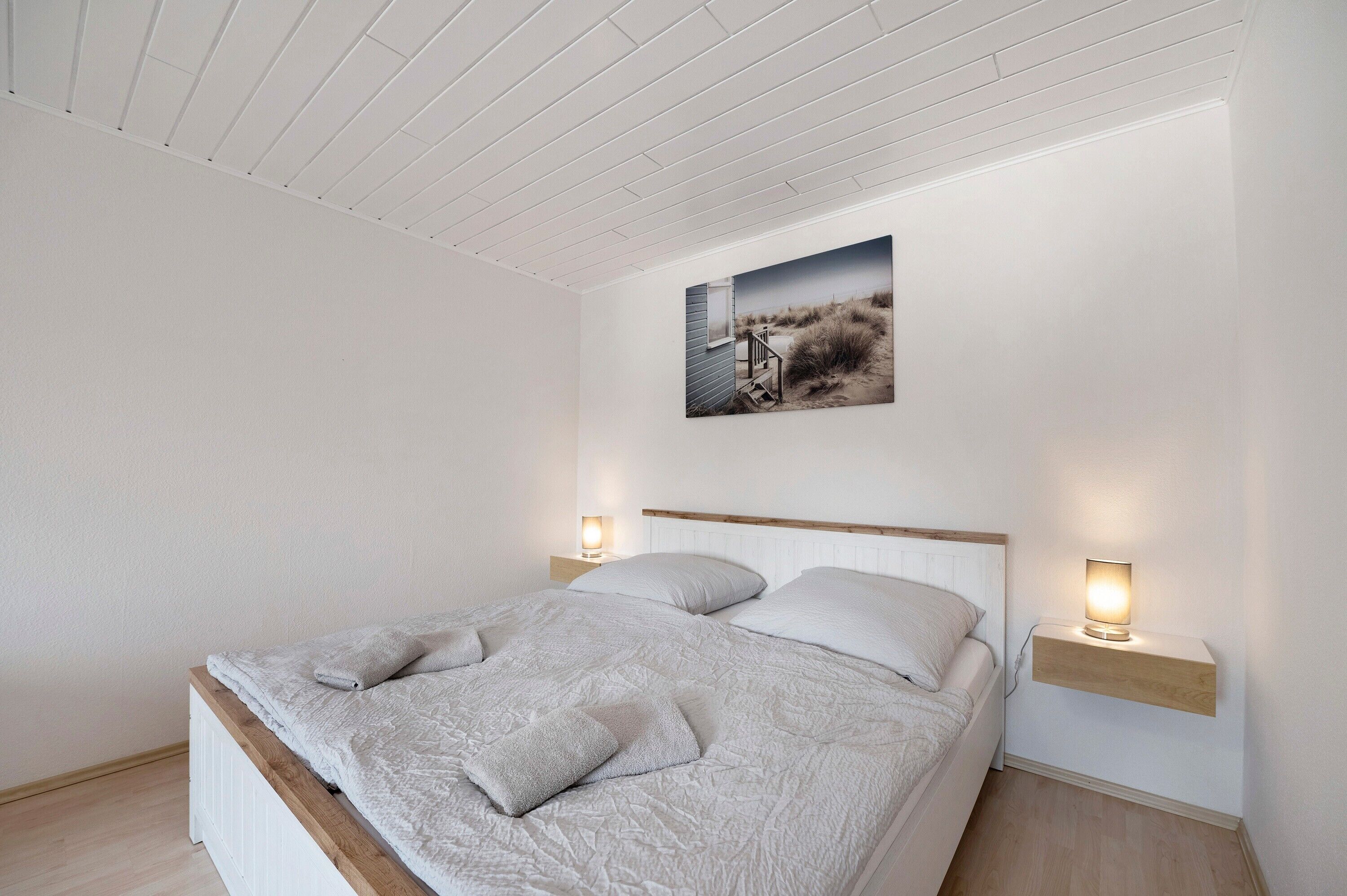 Un dormitorio con una cama, una mesita de noche con lámpara, un estante en la pared y un cuadro sobre la cama.