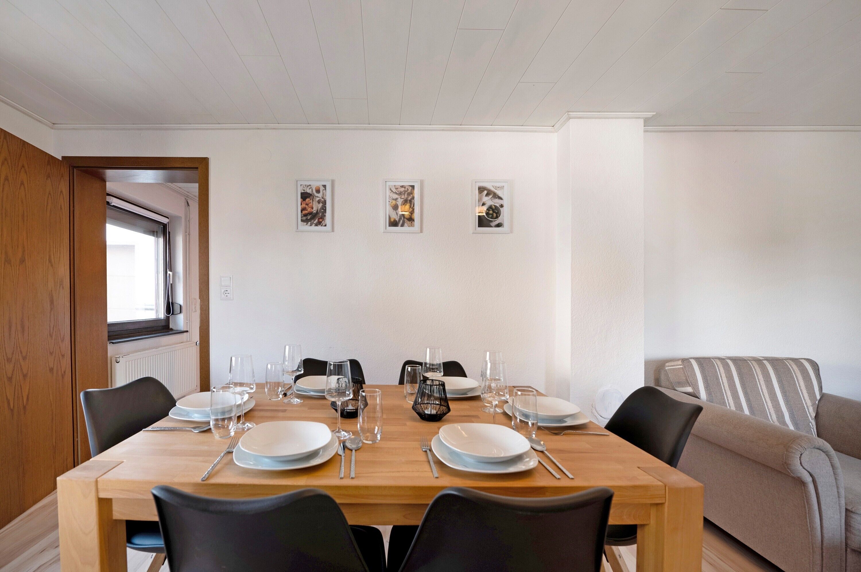 Un coin repas avec une table en bois dressée pour quatre personnes, des chaises et trois tableaux encadrés accrochés au mur.