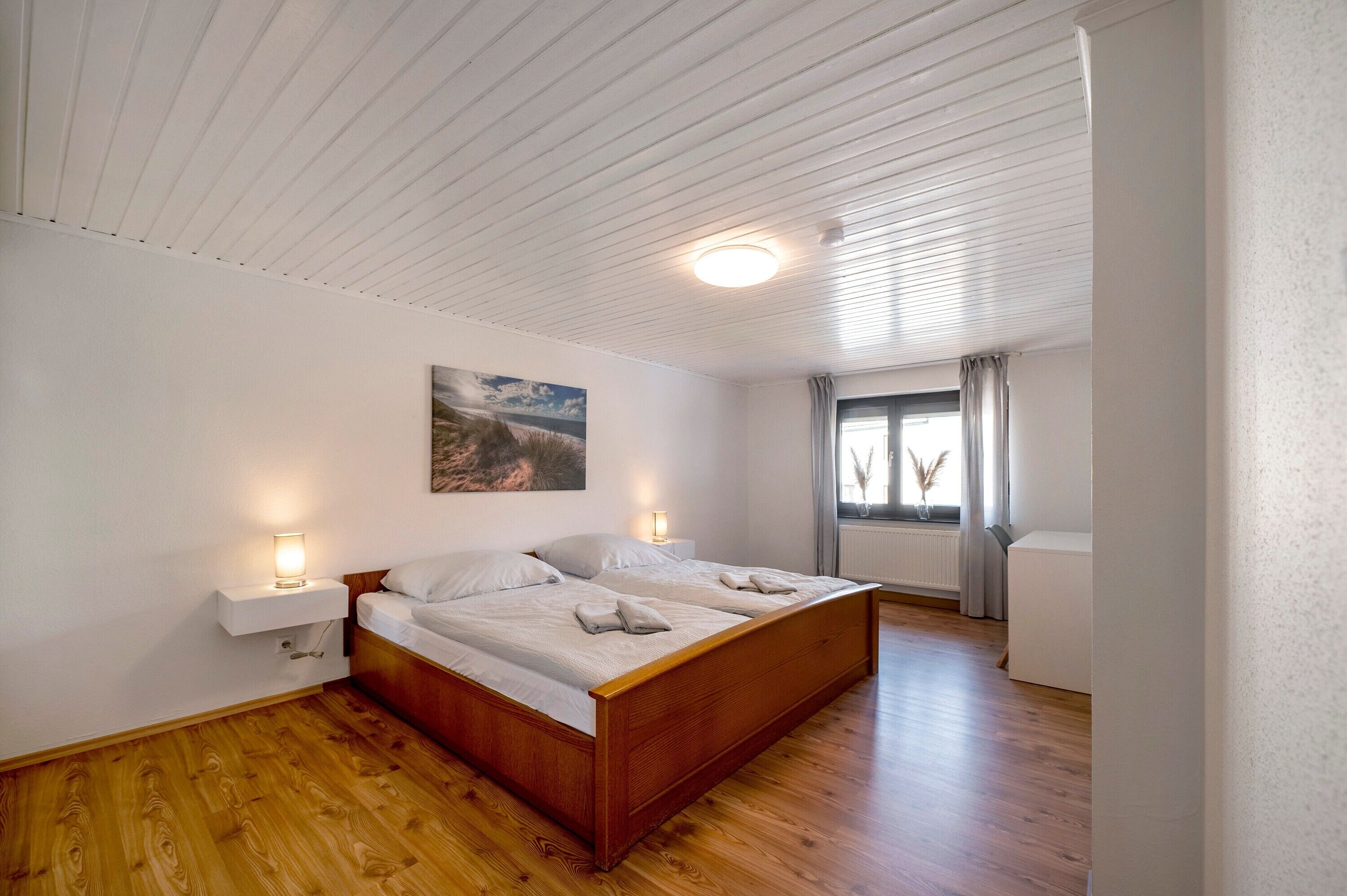 Een slaapkamer met een houten bed, witte wanden, een plafondlamp, een raam met gordijnen en een schilderij aan de muur.