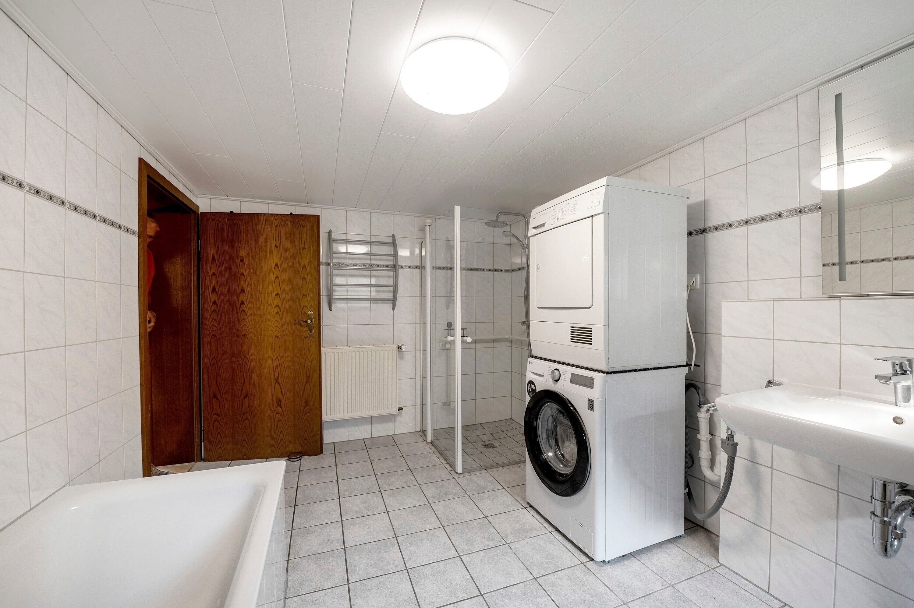 A bathroom with a white sink, a washing machine, a shower, and a wooden door.