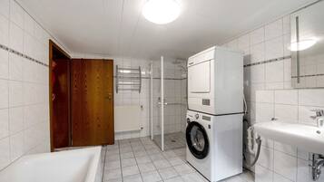 A bathroom with a white sink, a washing machine, a shower, and a wooden door.