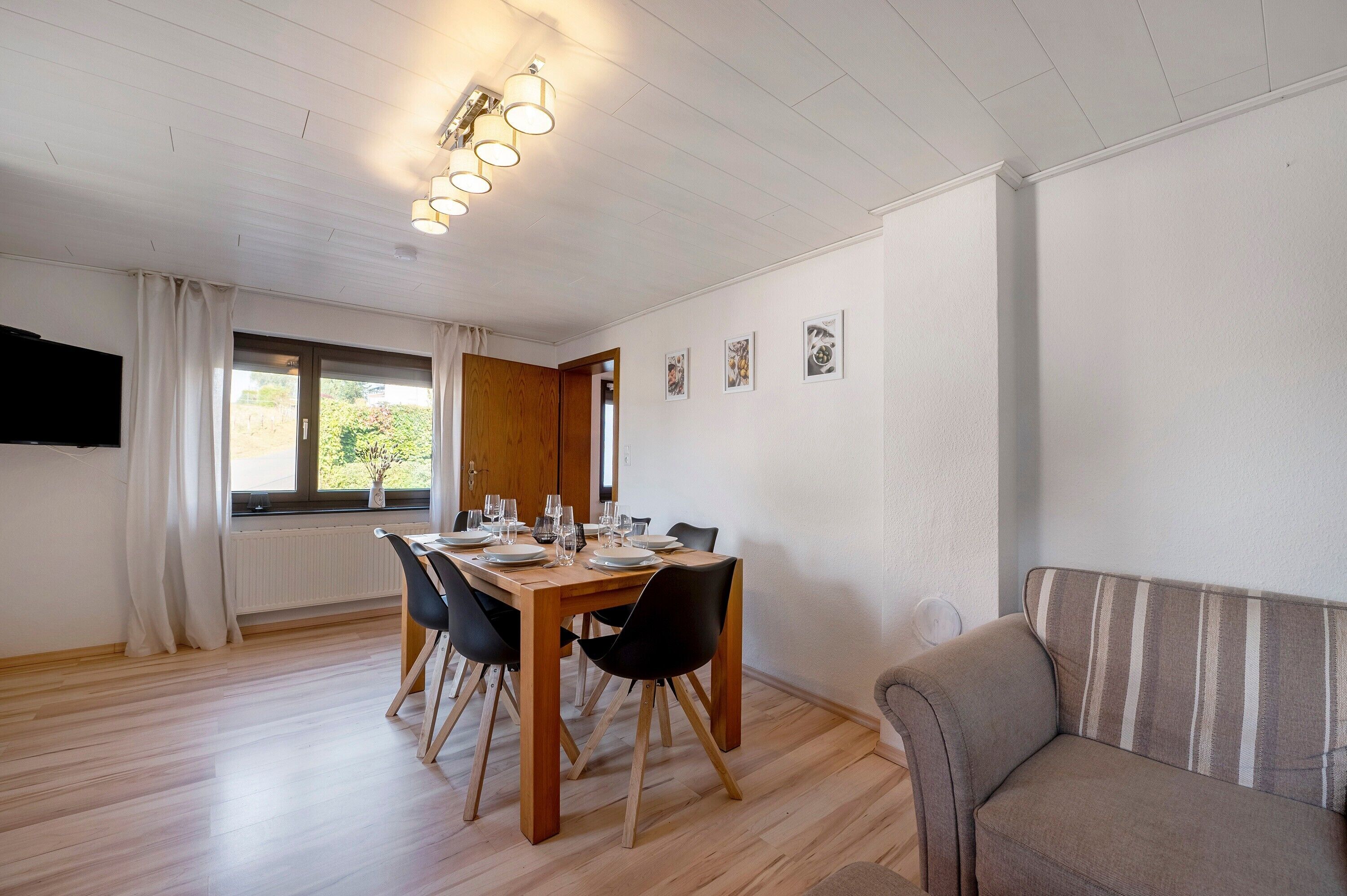 Un coin repas avec une table en bois dressée pour quatre personnes, des chaises noires, un canapé et un téléviseur fixé au mur.