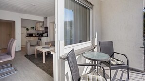 Un balcón con una mesa y sillas de vidrio, una puerta corrediza de vidrio que da a una cocina y vistas al exterior.