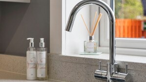 A modern kitchen with a stainless steel sink, a window with a view of greenery, and two bottles of hand soap on the countertop. - Bright and modern 2 bed flat with parking and a garden you can relax in (Edinburgh)