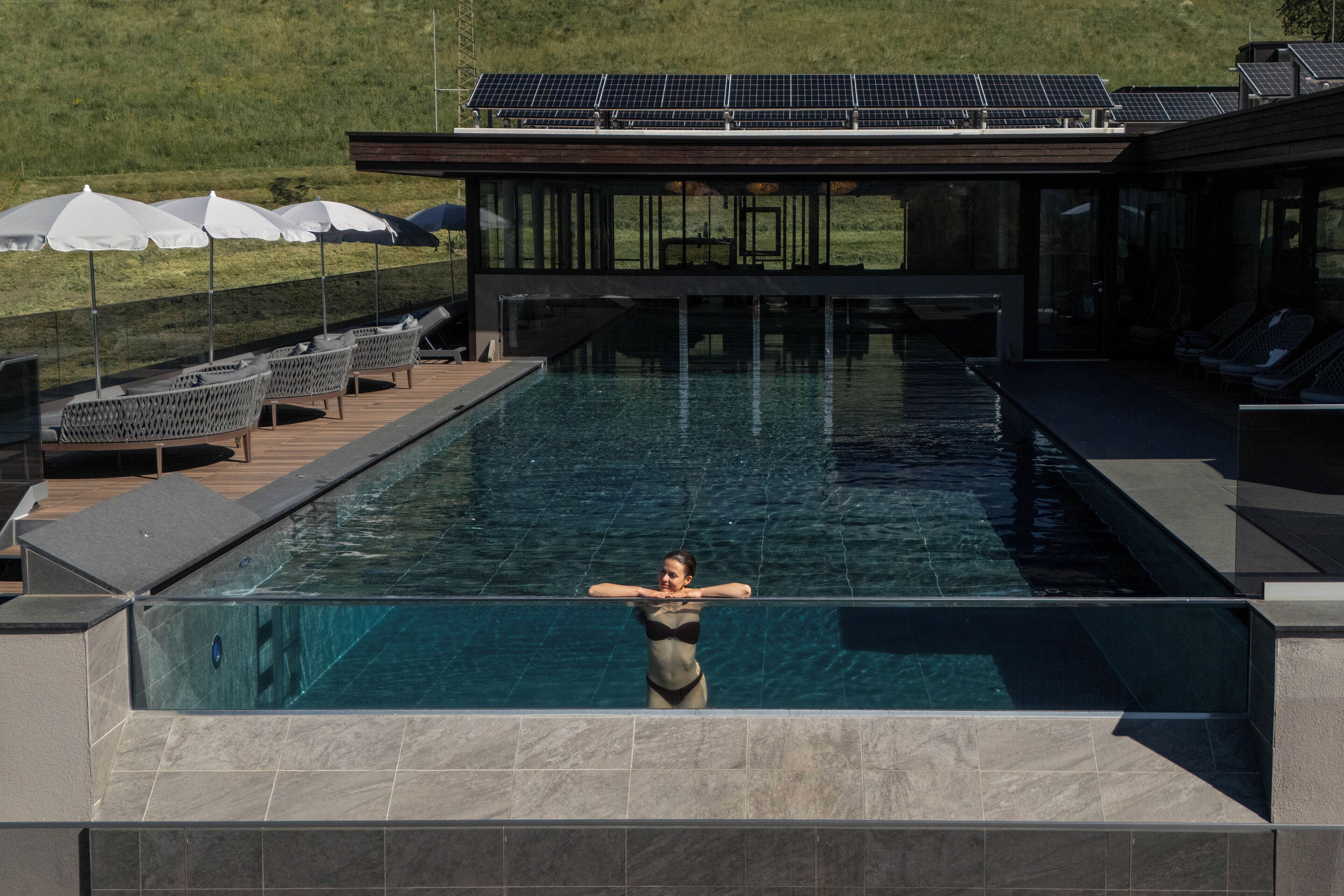 A swimming pool with a person in the water, lounge chairs, and a building with solar panels.