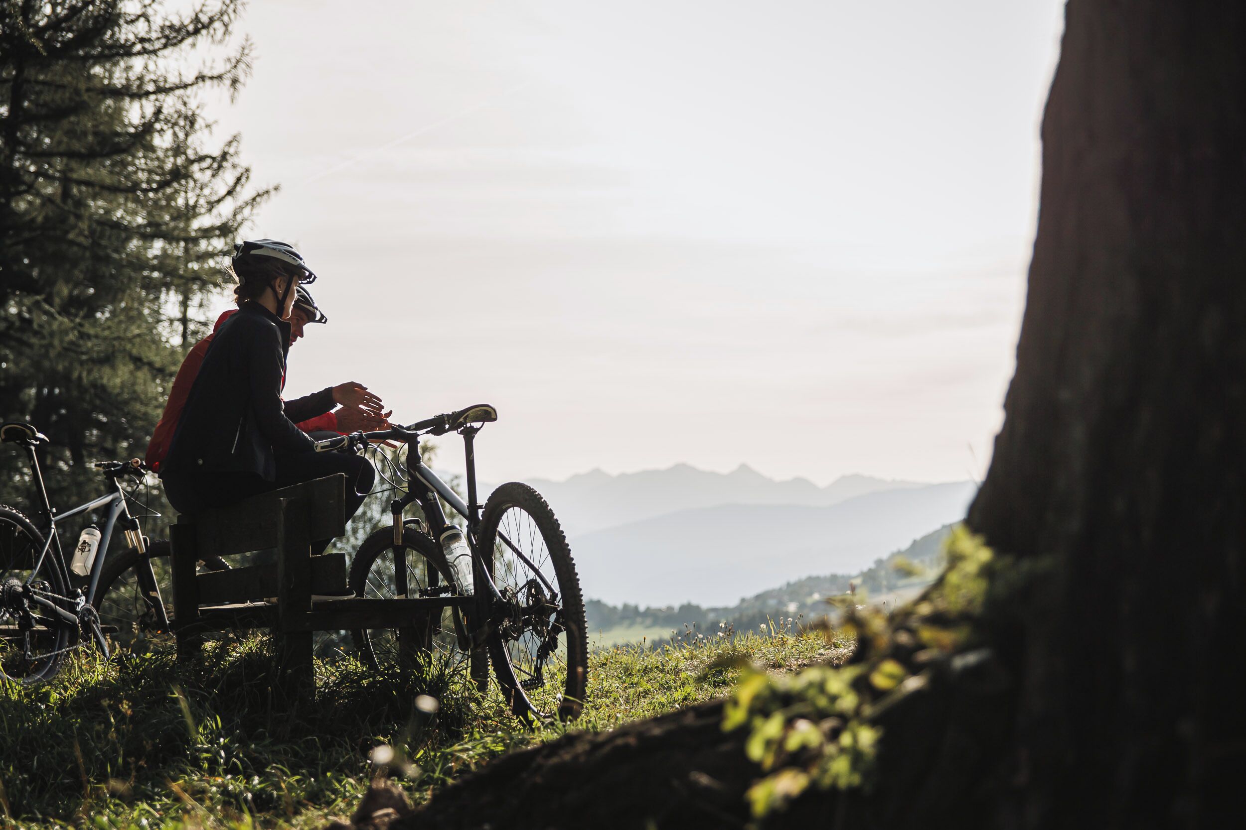 Una persona sentada en un banco con una bicicleta, rodeada de vegetación y montañas al fondo.