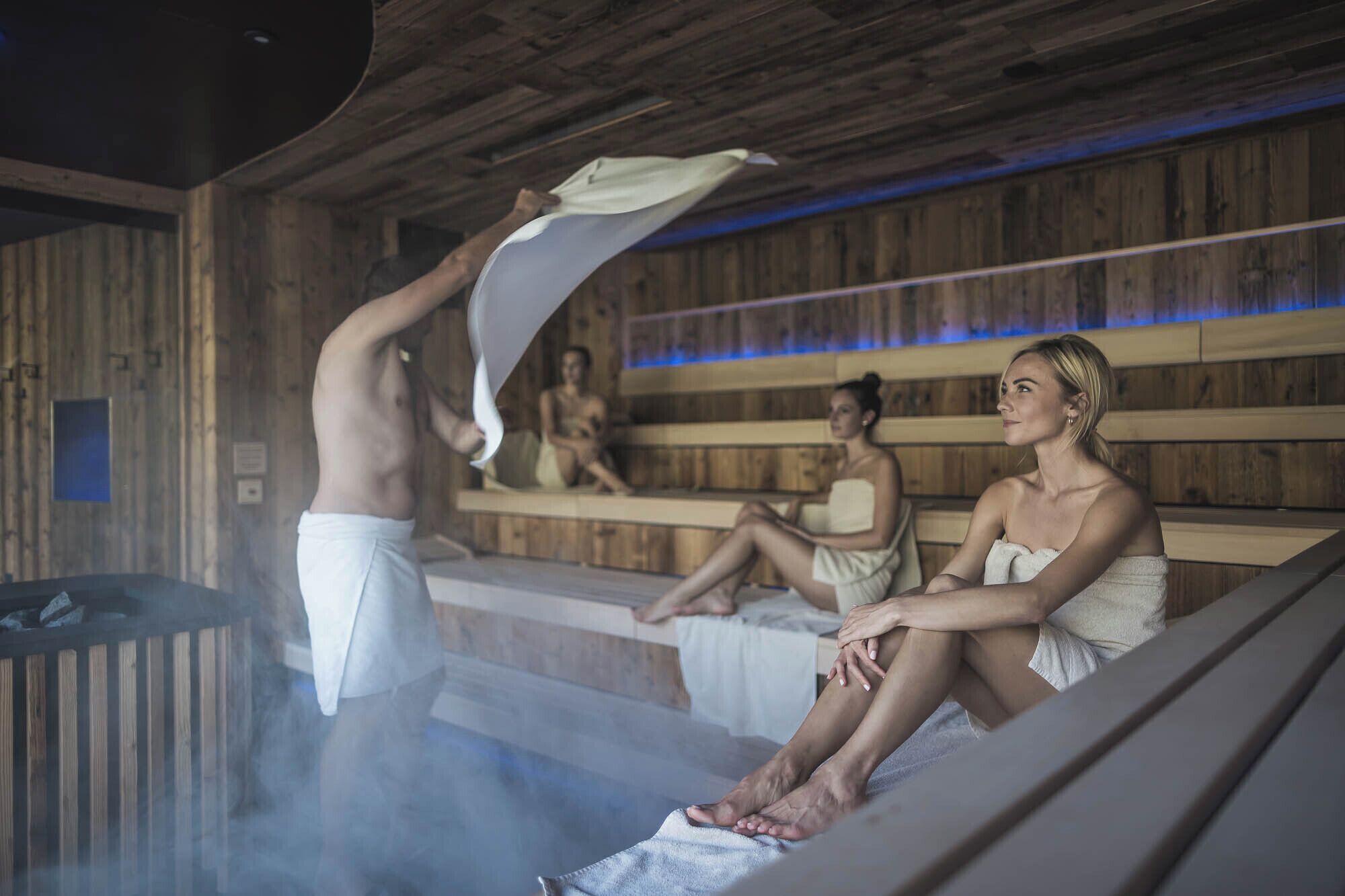 A sauna with wooden walls and benches, steam visible, and people relaxing.